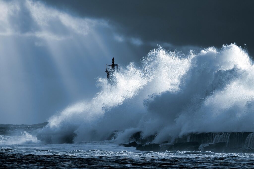 Massive ocean waves crashing over a small lighthouse during a storm, with light breaking through the clouds