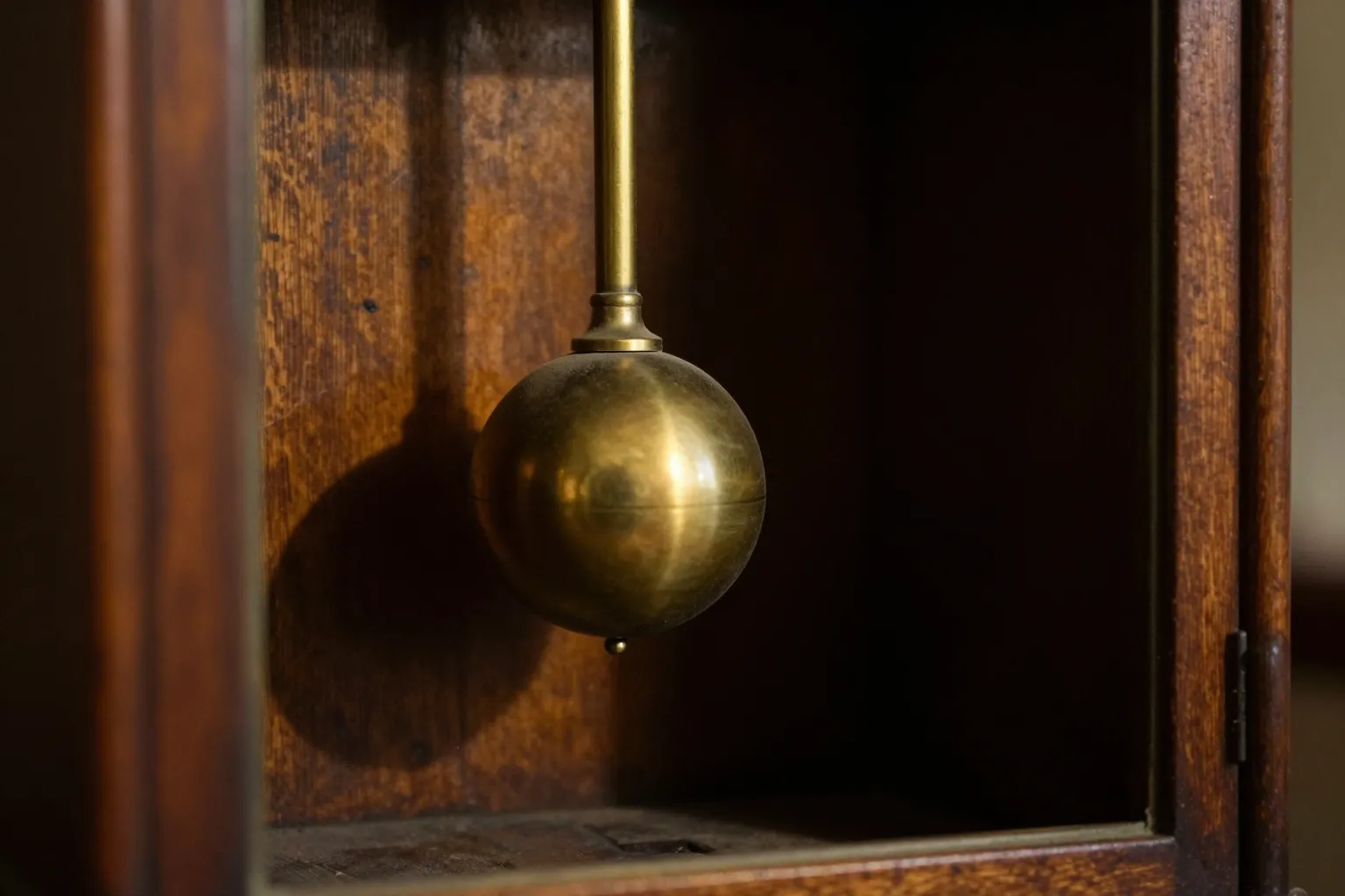 A heavy brass pendulum bob hanging inside a dark wooden clock case, its long shadow falling on the wall behind it