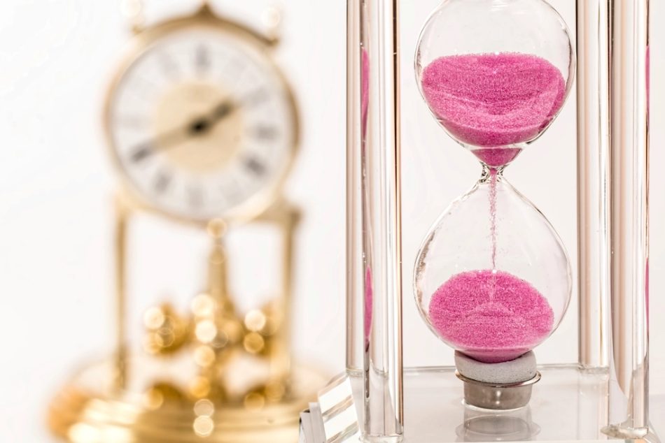 An hourglass with sand running through it, beside a brass mantel clock